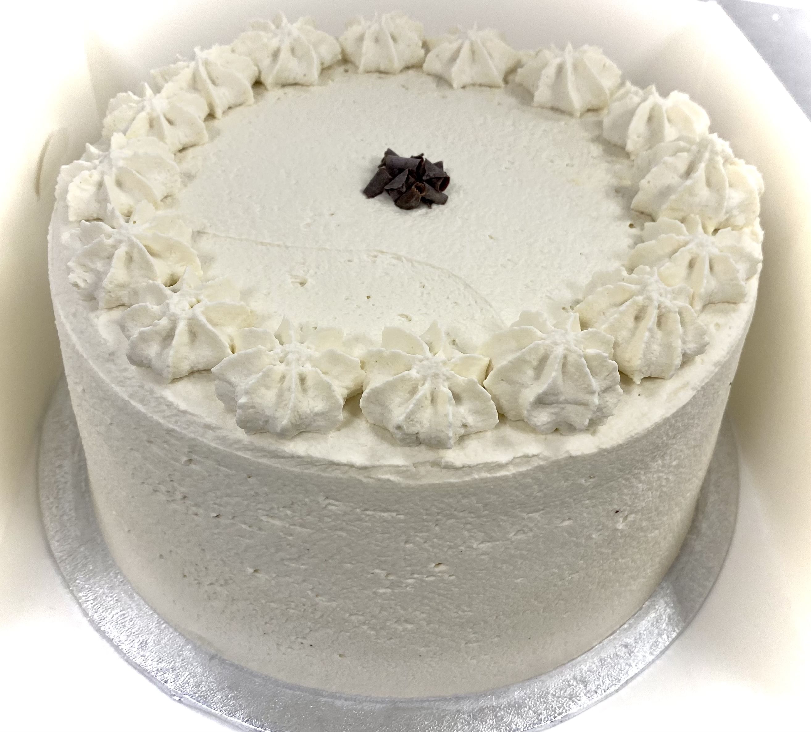 A round, white frosted cake with an even layer of smooth icing. The cake is topped with decorative dollops of white frosting along the edge and has a small cluster of dark chocolate shavings in the center. It is placed on a silver cake board.